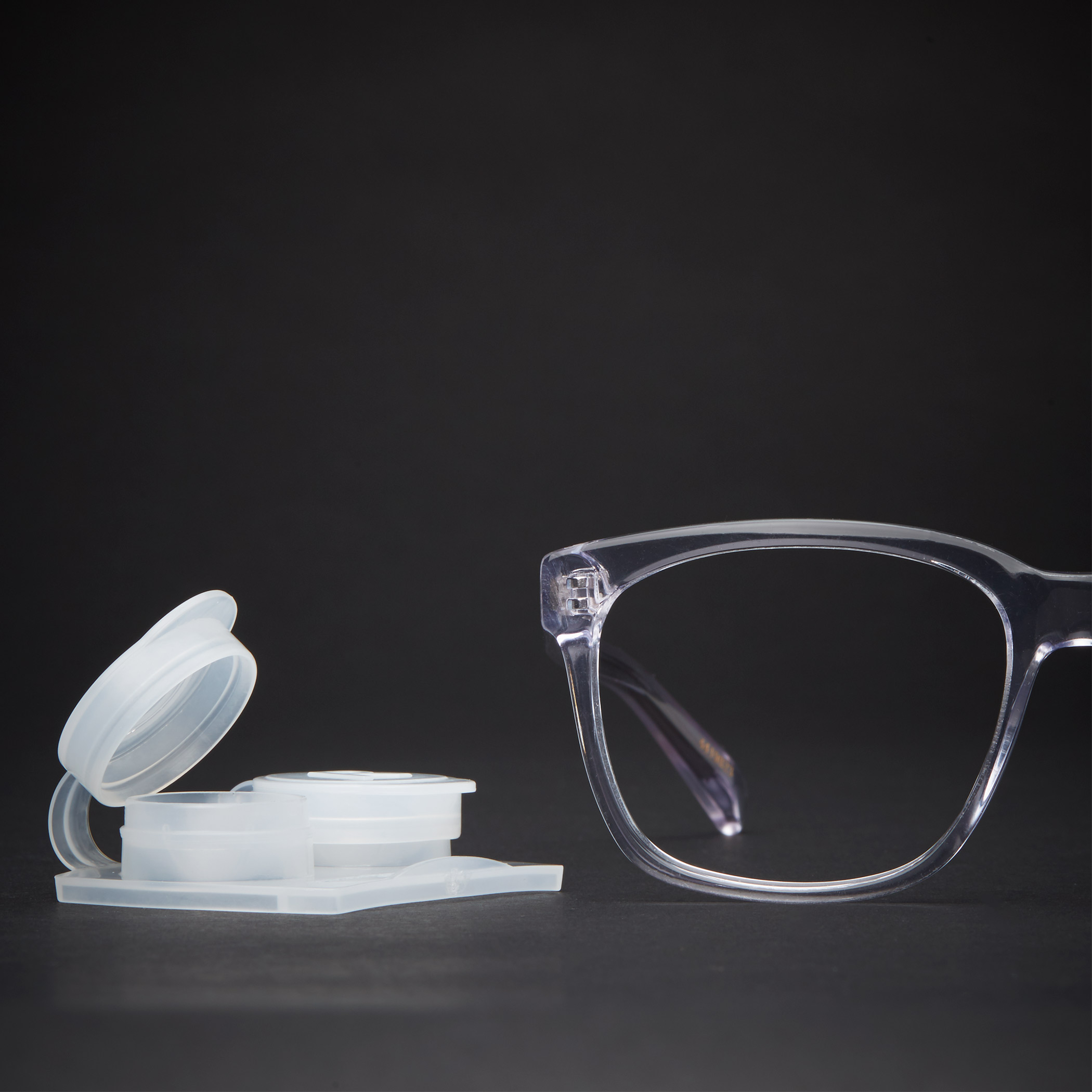 Clear eyeglasses next to an open white contact lens case on a black background.