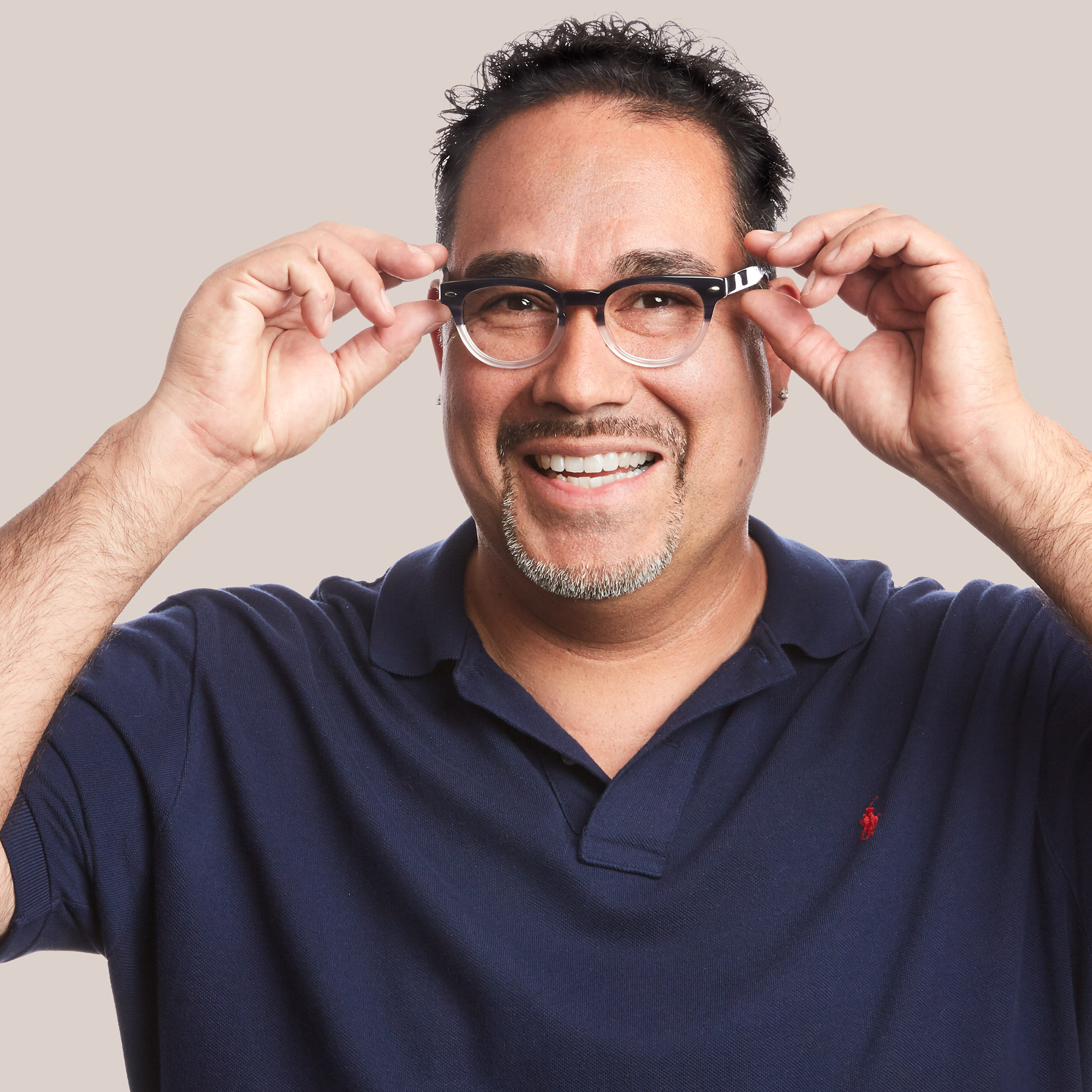 Man adjusting black-framed eyeglasses, wearing a navy blue collared shirt adorned with a small red logo.