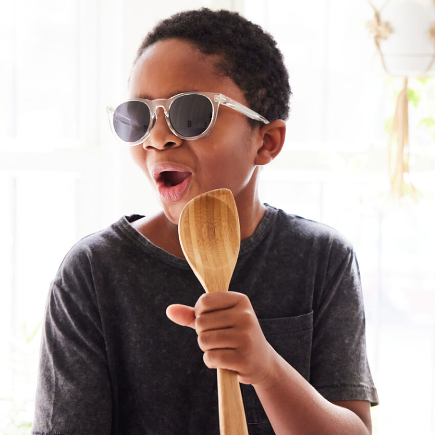Child singing while holding a wooden spoon, wearing sunglasses and a dark t-shirt.