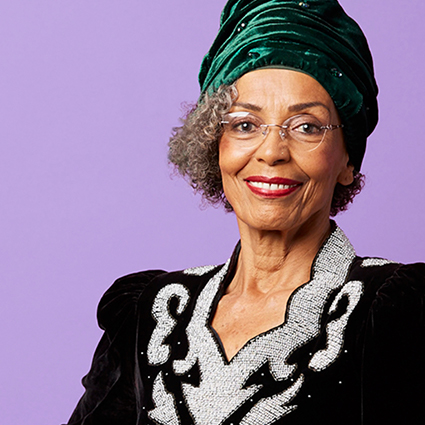 Elderly woman wearing a green turban, glasses, and a black outfit with silver embroidery detail.