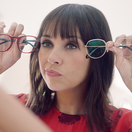 Woman trying on two different pairs of glasses, one with red frames and one with gold frames.