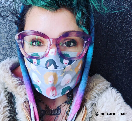 Person wearing a face mask with colorful cartoon faces, large purple glasses, and layered clothing. Text: @anna.arms.hair