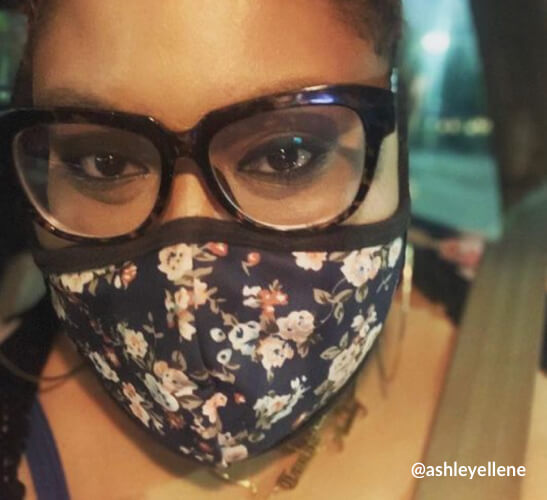 Person wearing large, dark-rimmed glasses and a dark floral face mask. Text: @ashleyellene