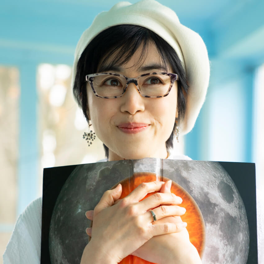 Smiling woman wearing glasses and a beret, holding a book open to an image of the moon and an orange sphere.