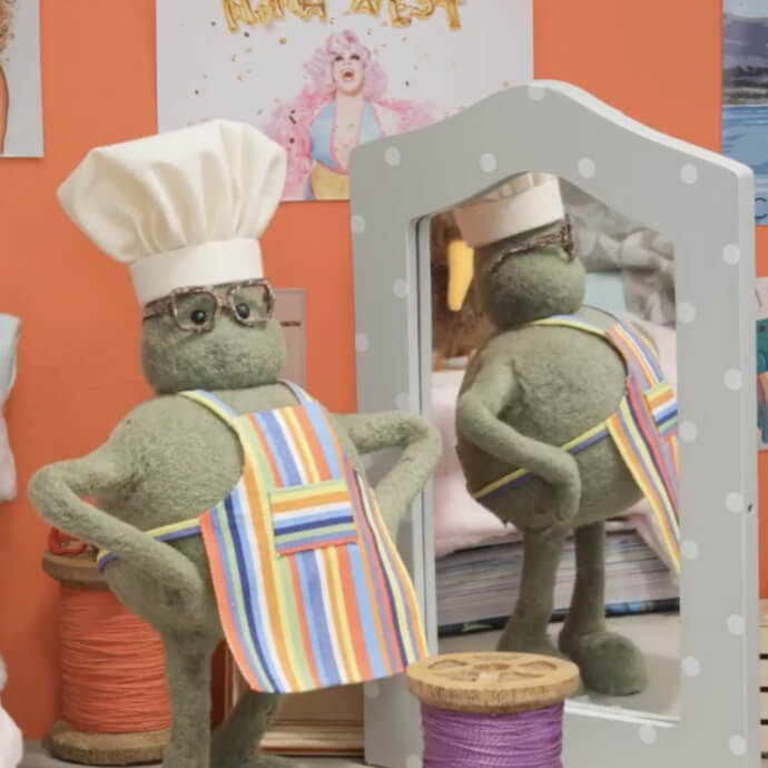 Green puppet wearing chef hat and striped apron standing confidently in front of a mirror.