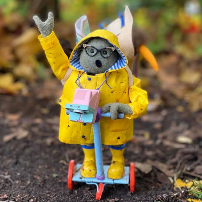 Plush toy in a yellow raincoat on a blue scooter, wearing glasses and a backpack.