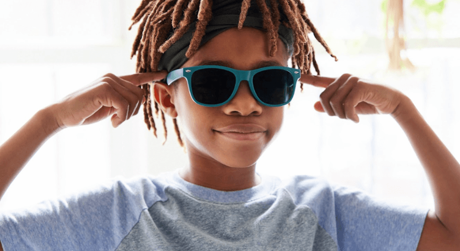 Child wearing blue sunglasses and light blue T-shirt, pointing at their sunglasses with both index fingers.