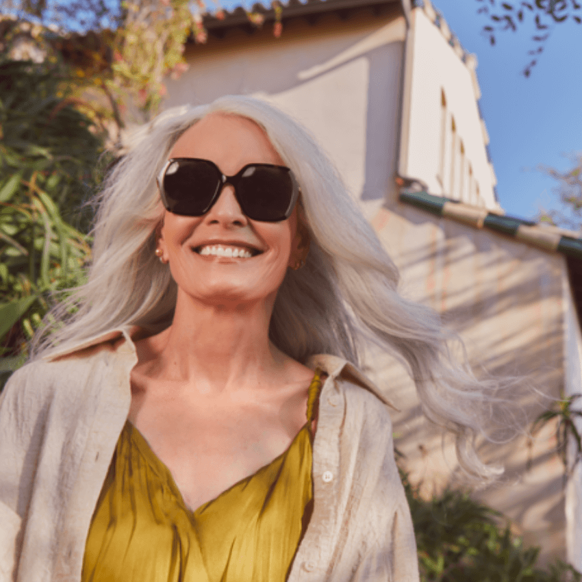 Woman wearing large black sunglasses, a green dress, and a beige open shirt smiling outdoors.