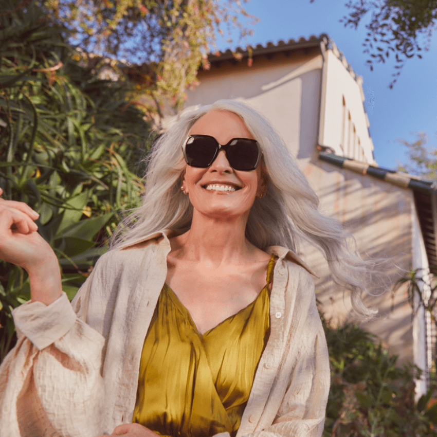 Woman in golden dress wearing black sunglasses and a light beige shirt, smiling confidently.
