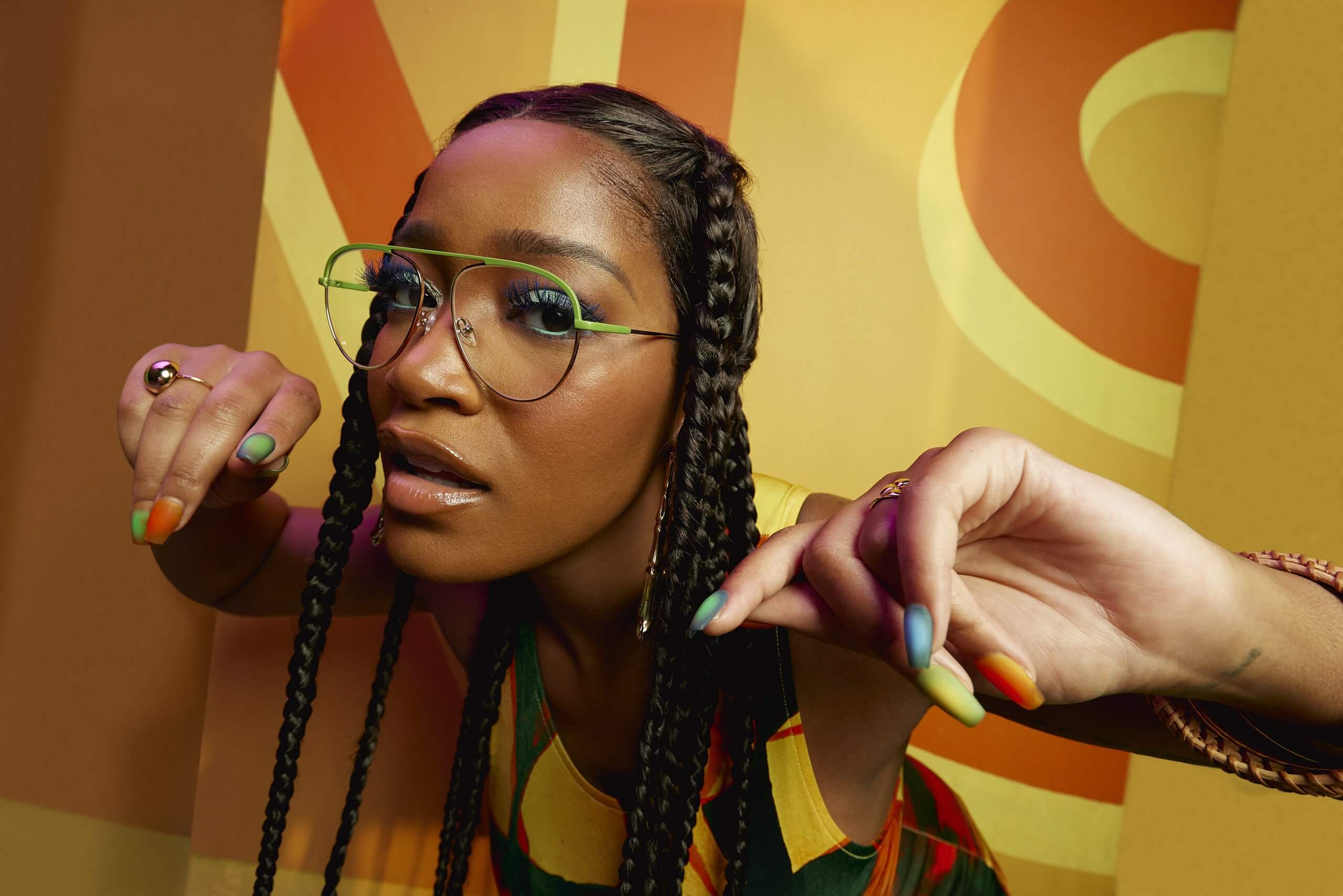 Woman with green-frame eyeglasses and braids, wearing colorful nail polish and a vibrant patterned top.