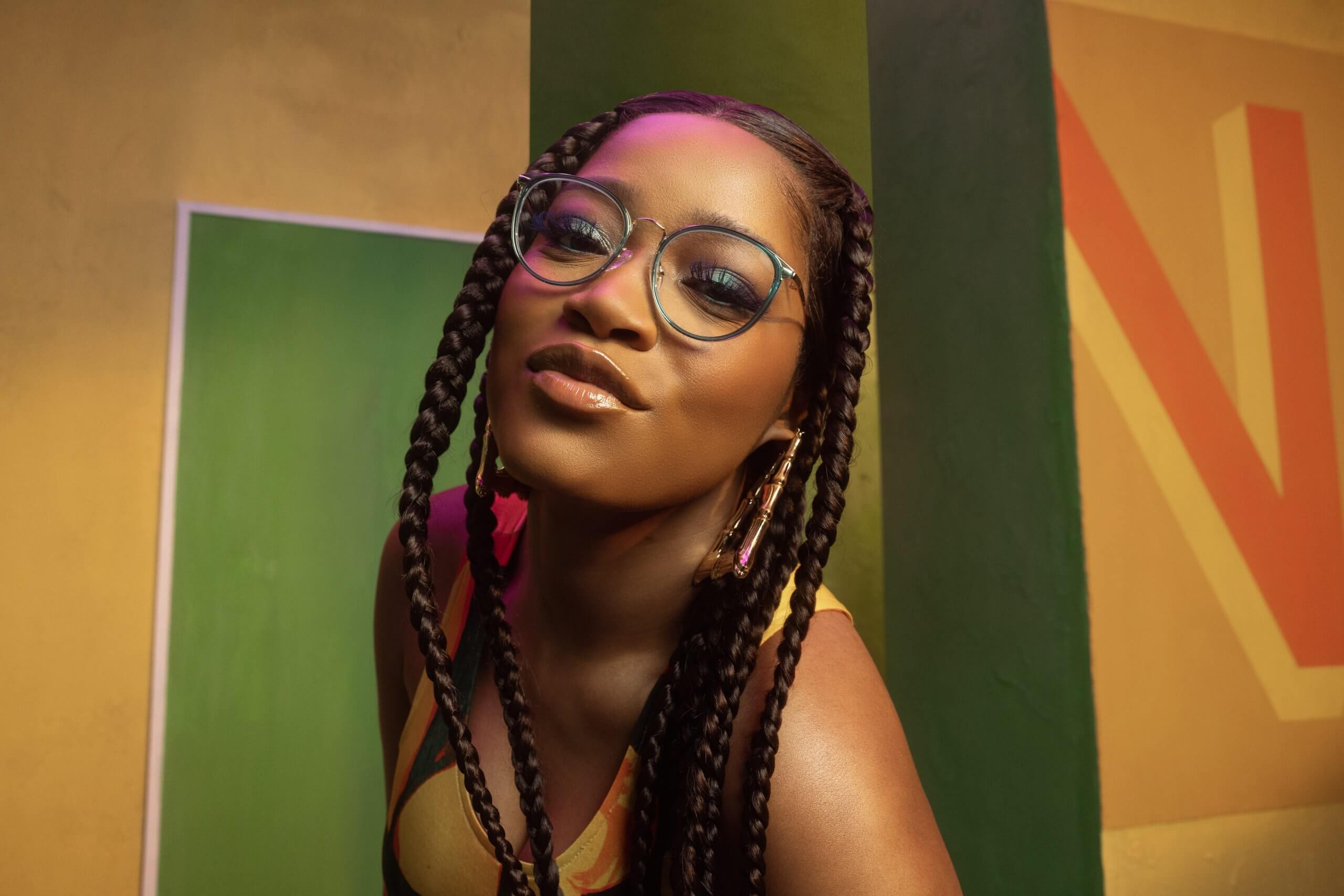 Person wearing round glasses with braided hair and hoop earrings, posing with a confident expression.
