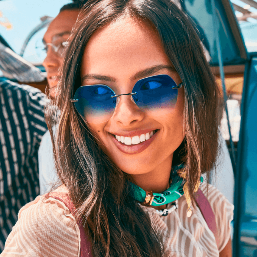 Woman wearing blue gradient sunglasses and a green scarf, smiling in an outdoor setting.