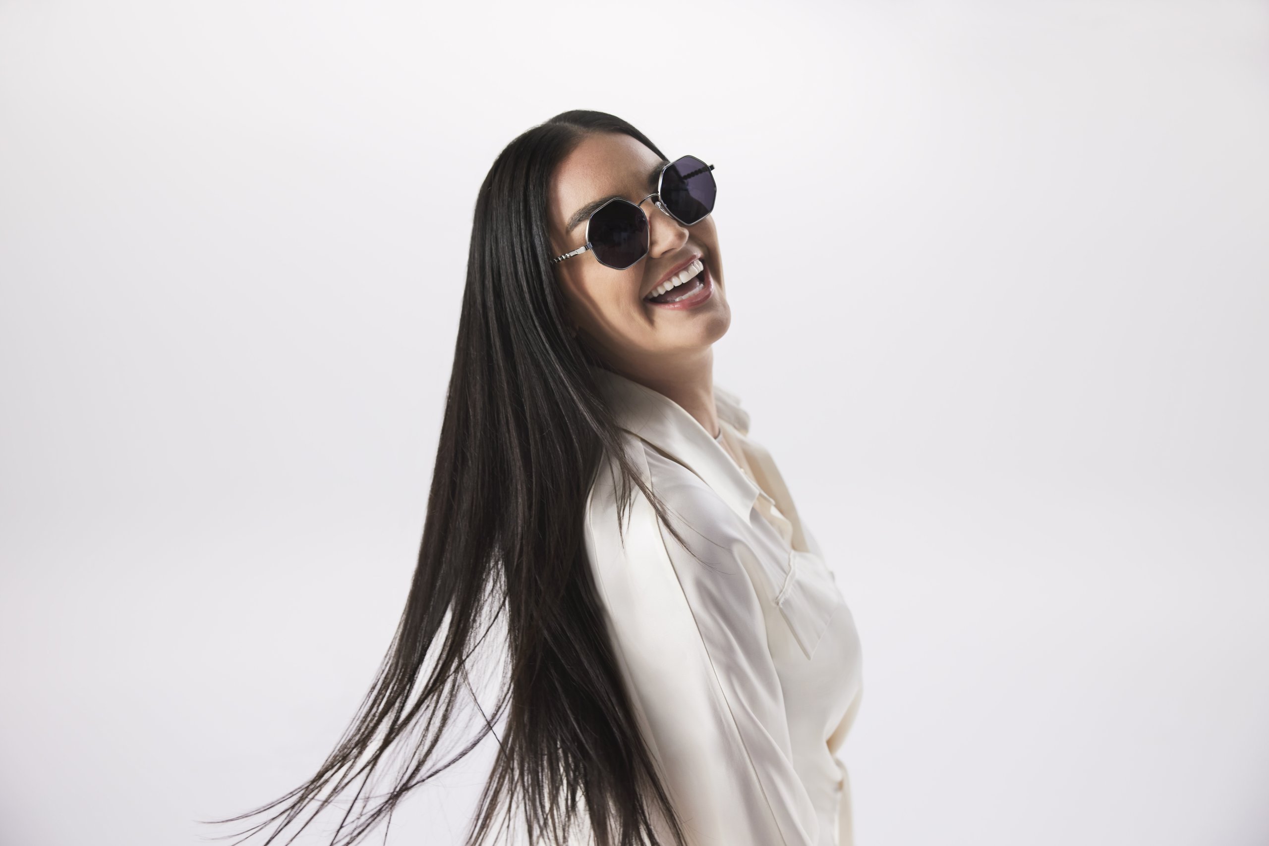 Woman wearing round, dark sunglasses and a white shirt, smiling and flipping her long, straight hair.