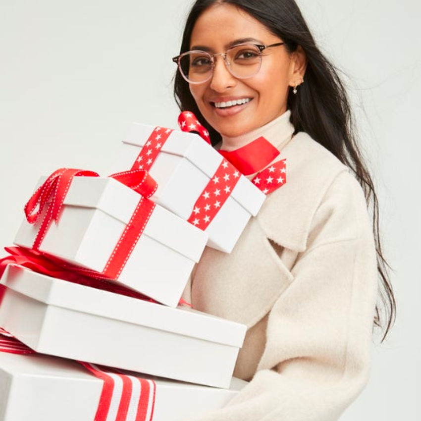 Woman holding white gift boxes with red ribbons and stars, in beige coat and glasses, smiling.