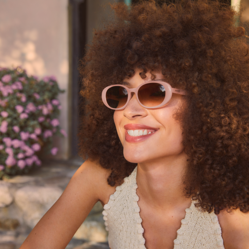 Woman with curly hair wearing pink oval sunglasses, smiling brightly in a sleeveless knit top.