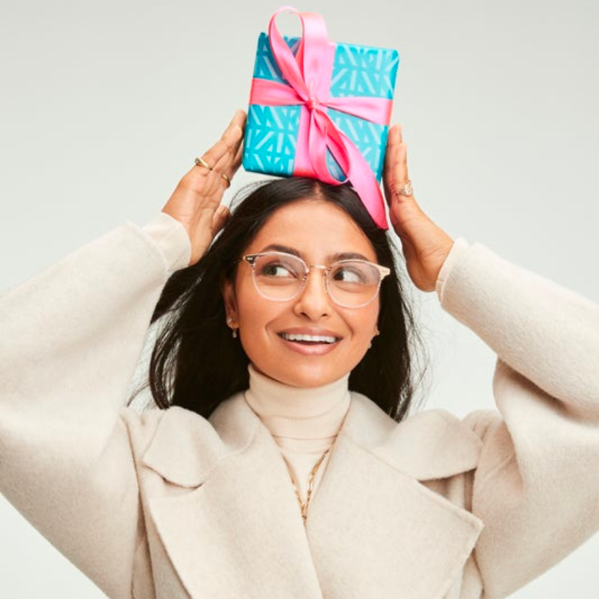 Woman holding a blue gift box with a pink ribbon on her head, wearing a beige coat and eyeglasses.