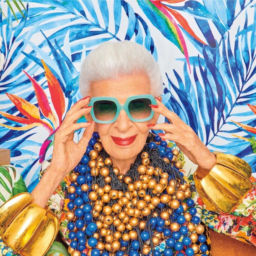 Woman wearing large turquoise sunglasses, multiple bead necklaces, and oversized gold cuff bracelets.