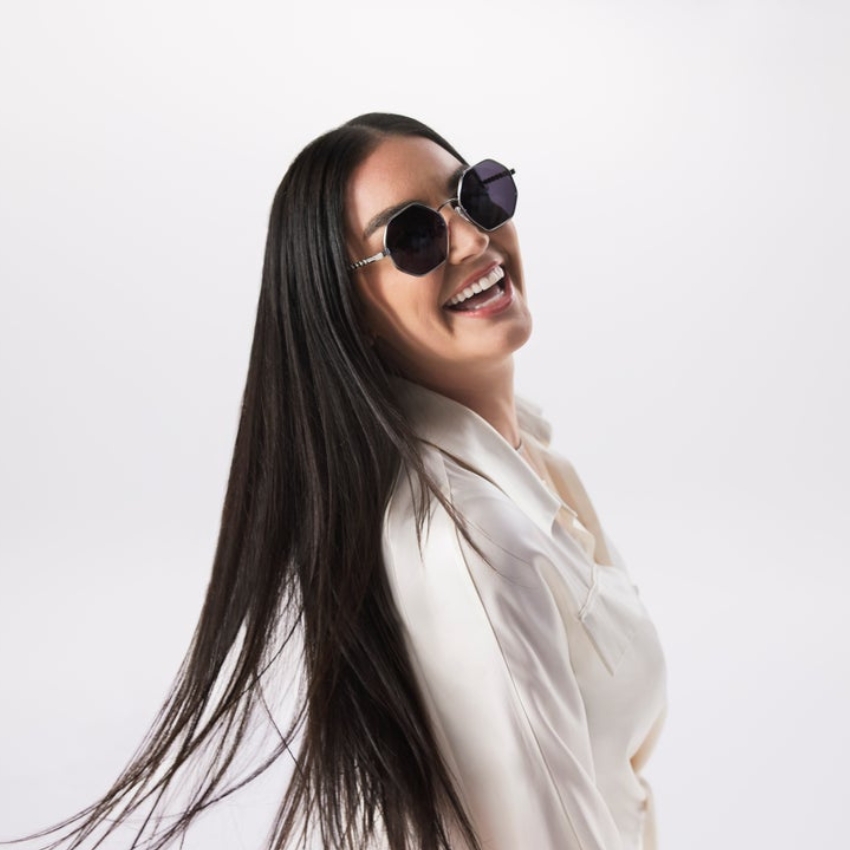 Woman wearing black, round sunglasses, smiling, and looking over her shoulder. Long hair flows down.