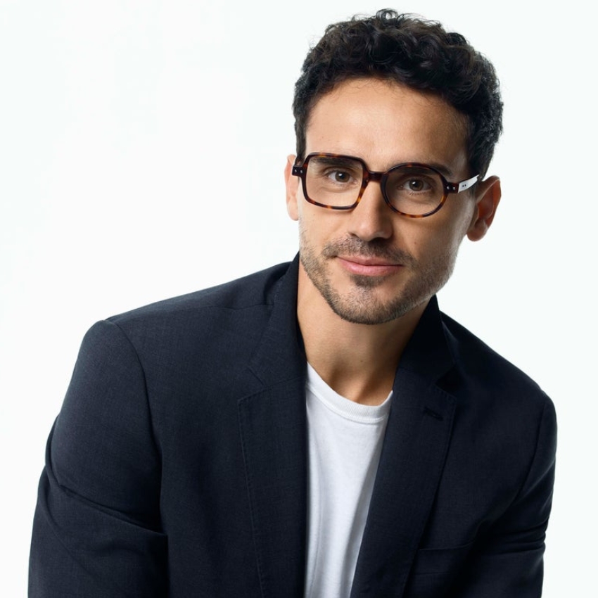 Man wearing brown tortoiseshell eyeglasses, a black blazer, and a white t-shirt.