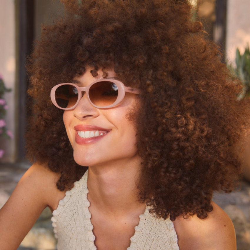 Woman wearing light pink oval sunglasses with brown-tinted lenses, smiling outdoors.