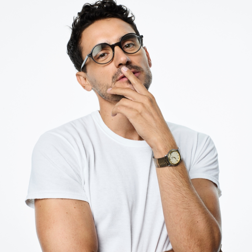 Man wearing a white T-shirt, black-rimmed eyeglasses, and a gold wristwatch with a contemplative expression.