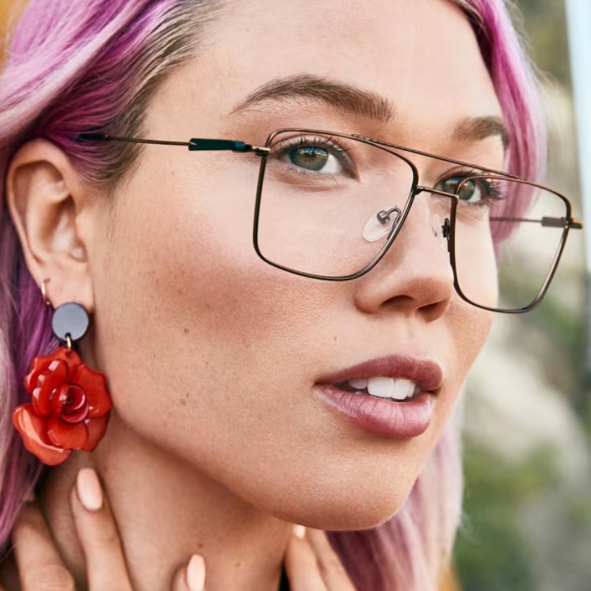 Wearing oversized square glasses with metal frames, red flower earring on the left ear, pink dyed hair
