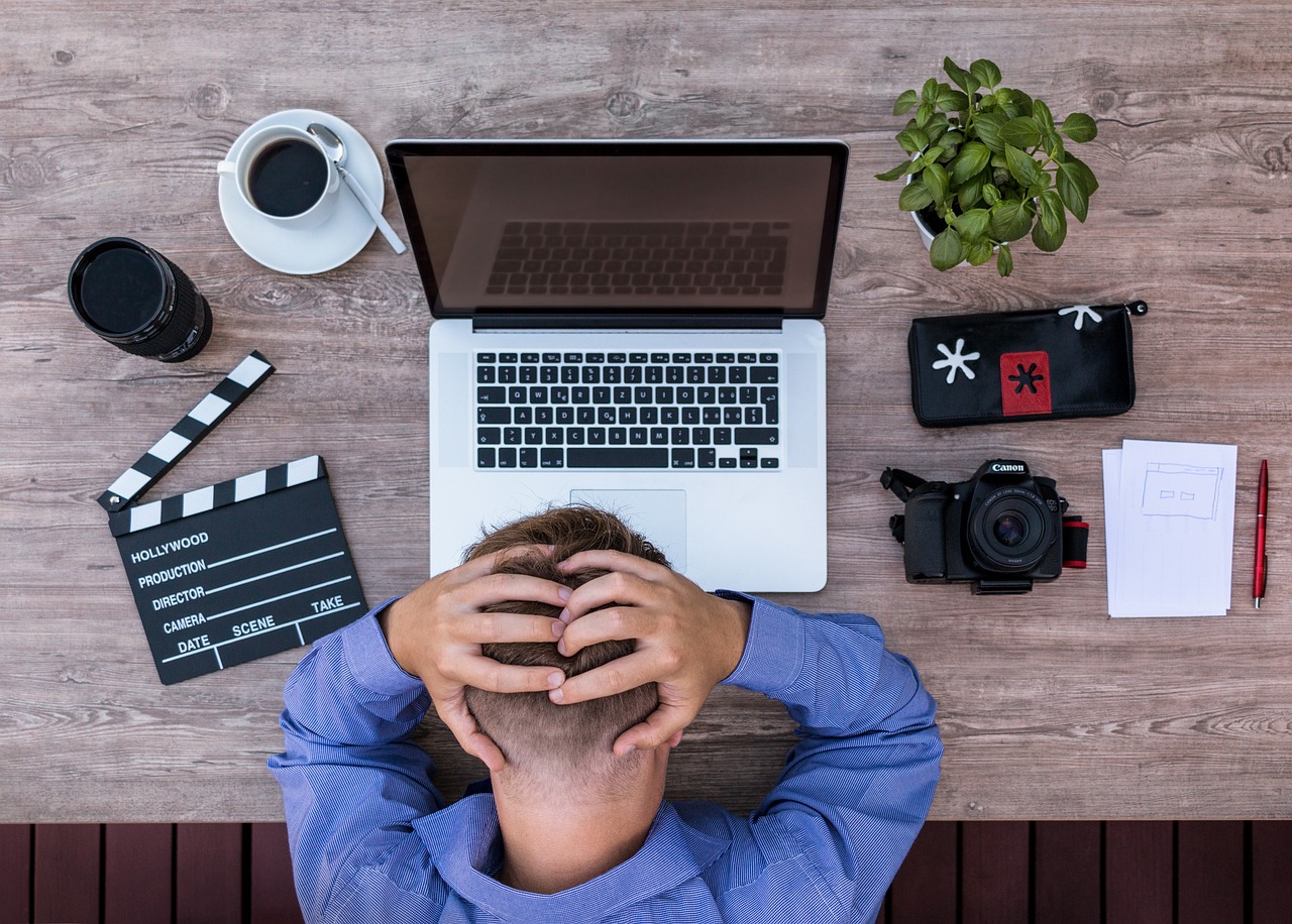 Man holding head in front of open laptop; nearby items include camera, coffee cup, and clapperboard. Text on clapperboard: "HOLLYWOOD, PRODUCTION, DIRECTOR, CAMERA, DATE, SCENE, TAKE".