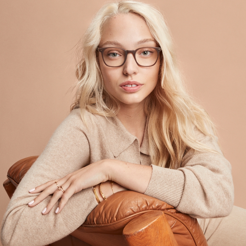 Woman wearing rectangular eyeglasses with a light brown frame, posing with an arm resting on a leather chair.