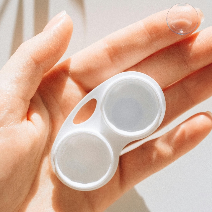 White contact lens case held in hand, with a single contact lens resting on fingertip.