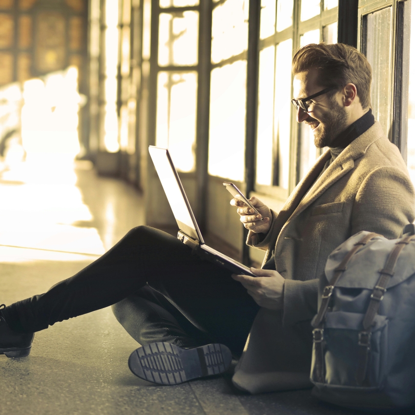 Man using a laptop and smartphone seated on the floor with a backpack beside him.