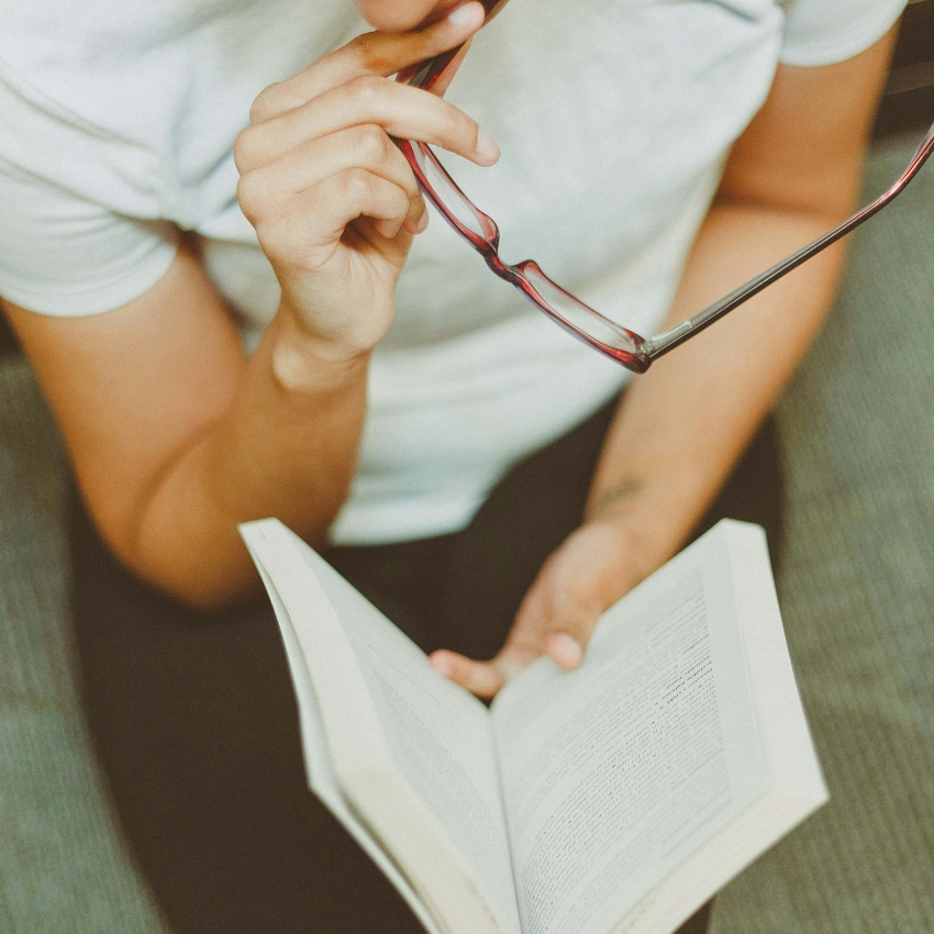 Holding a book while pondering with glasses in hand.