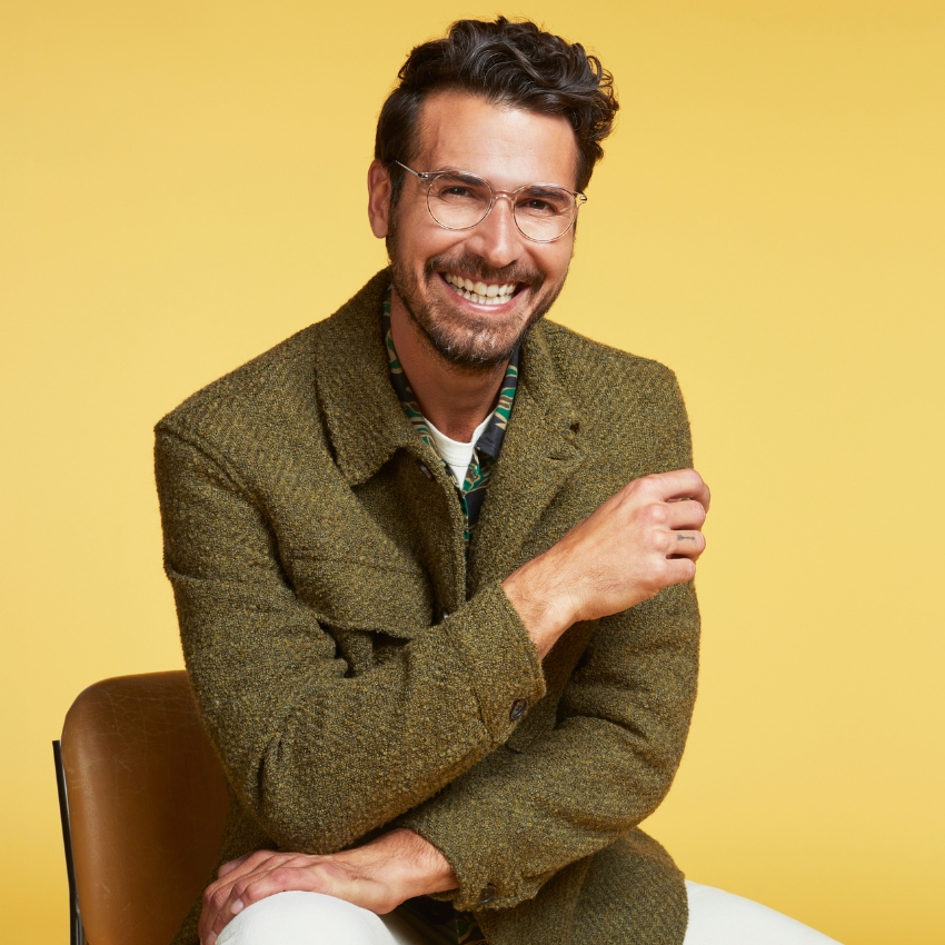 Smiling man wearing a green tweed jacket and clear eyeglasses against a yellow background.
