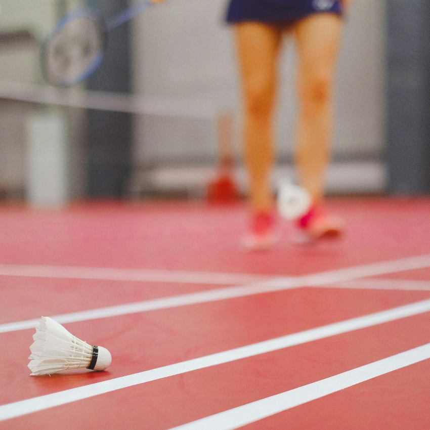 Badminton shuttlecock on a red court with white lines. Player in the background holding a racket, blurred.