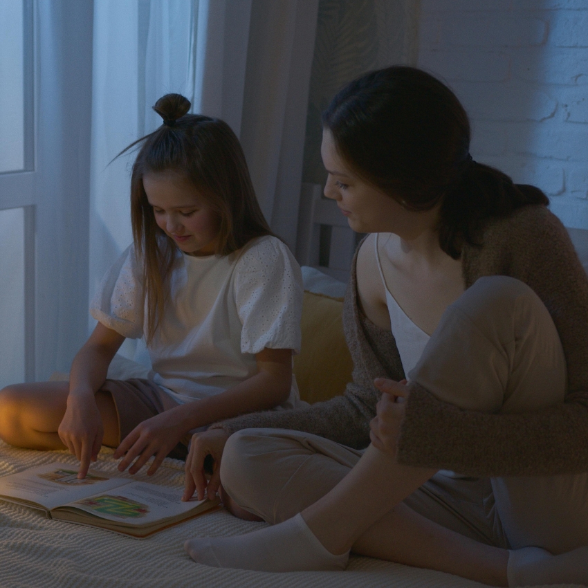 Child and adult reading a picture book together on a bed.