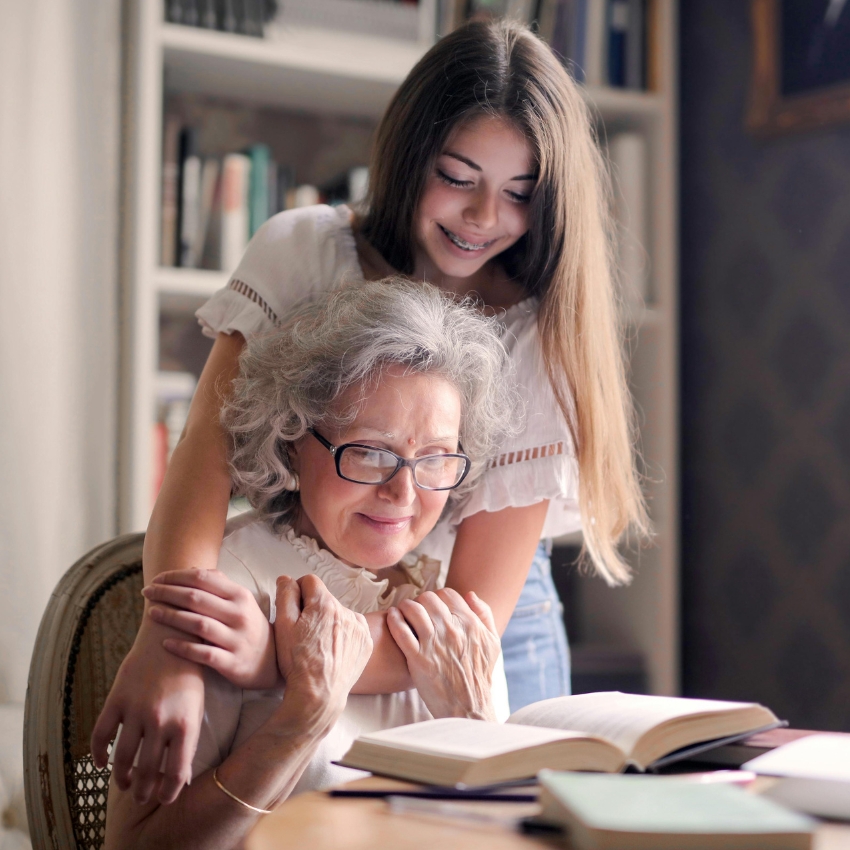 Older woman wearing glasses reading a book, while younger woman in white top embraces her from behind.