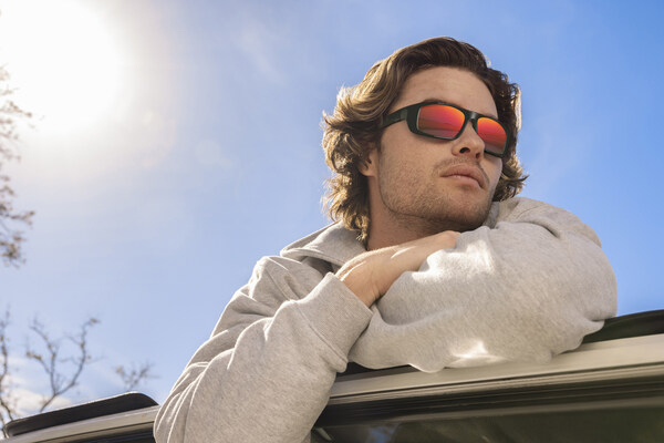 Man wearing sunglasses with red reflective lenses leaning on car.